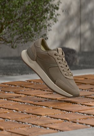 Zapatilla deportiva beige informal con paneles texturizados y lisos, cordones y suela blanca flotando sobre suelo exterior de baldosas con planta verde de fondo.