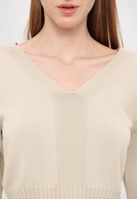 Maglione beige lavorato a maglia con scollo a V, orlo a coste e dettagli in pannelli testurizzati. Il tessuto ha una consistenza liscia e un aspetto leggero.
