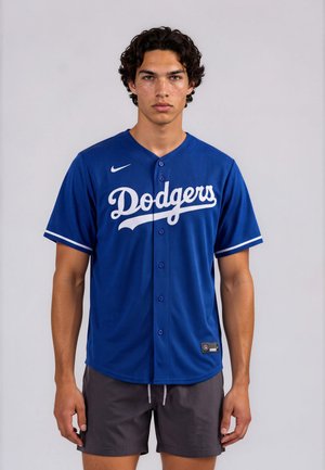 Junger Mann mit lockigem Haar, der ein blaues Dodgers-Baseballtrikot und dunkle Shorts trägt, vor einem schlichten hellen Hintergrund.
