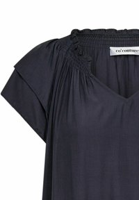 Tumma harmaa paita, joka on valmistettu kevyestä kankaasta. Siinä on V-pääntie, rypistetty smokkityyli pääntien ympärillä ja lyhyet hihat.