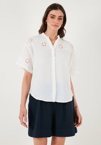White button-up shirt with short sleeves, featuring floral cut-out embroidery on shoulders, made of lightweight fabric. Paired with navy shorts.