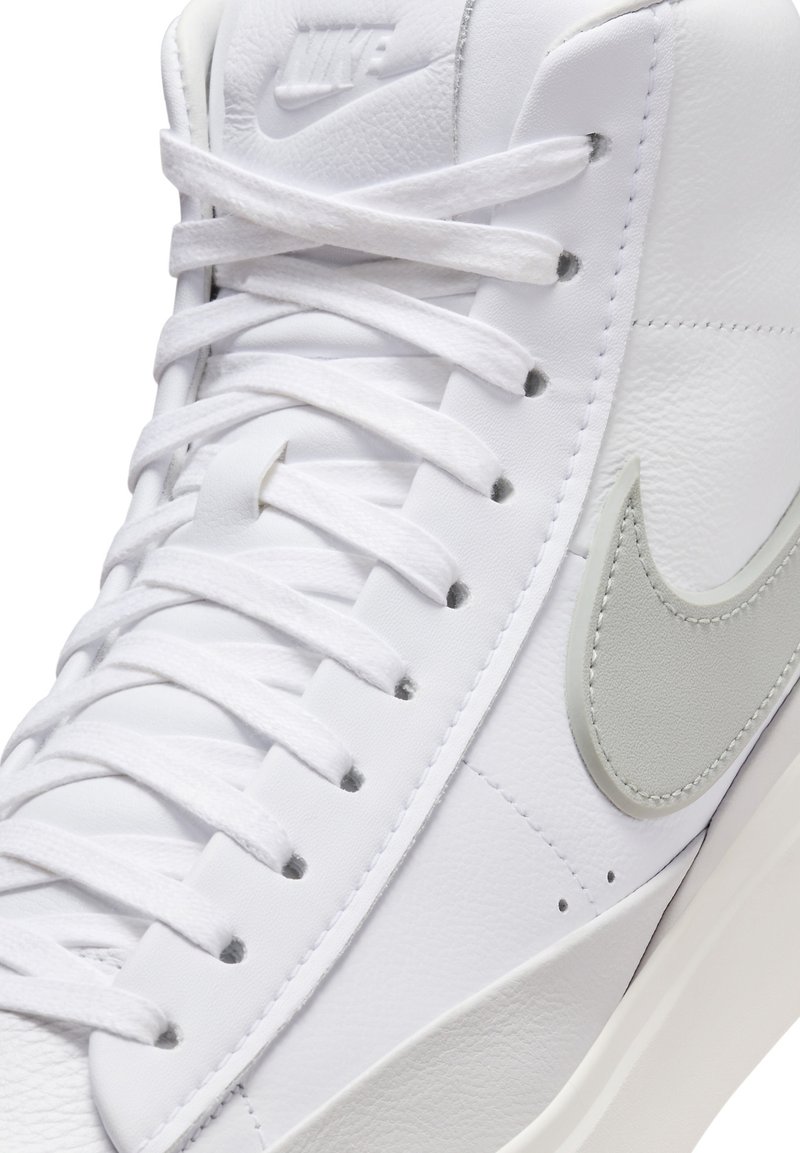 Sneaker bianca realizzata in pelle testurizzata, con il logo Nike grigio chiaro, lacci bianchi piatti e una suola in gomma liscia. Vista laterale con cuciture intricate.