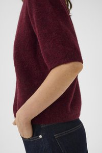 Personne portant un pull en fausse fourrure bordeaux à manches courtes, avec une main glissée dans la poche d'un jeans en denim bleu foncé.
