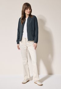 Mörkblå lätt bomberjacka med elastiska manschetter, kombinerad med en vit skjorta och krämfärgade byxor, bärs med beige sneakers.