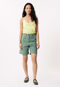Korte groene gestreepte shorts, gecombineerd met een lichte, mouwloze, glanzende gele top. Het schoeisel heeft groene accenten en een gesp.
