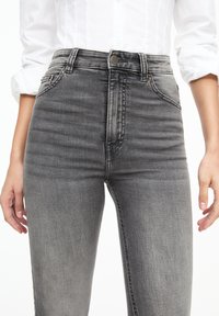 Mujer vestida con jeans ajustados de tiro alto en gris deslavado y una camisa blanca de botones con las mangas arremangadas, con las manos relajadas a los lados.