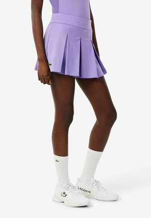 Femme portant une jupe de tennis plissée violette, des chaussettes blanches montantes avec un petit logo, et des baskets blanches Lacoste sur un fond uni.