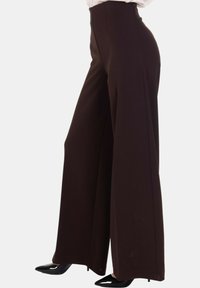 Pantaloni a vita alta, a gamba larga di colore marrone scuro indossati con tacchi neri a punta, mostrati dalla vita ai piedi su uno sfondo bianco.