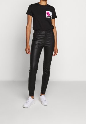 Persoon in zwart t-shirt met roze graphic, zwarte leren broek en witte sneakers, staand tegen een effen lichte achtergrond.