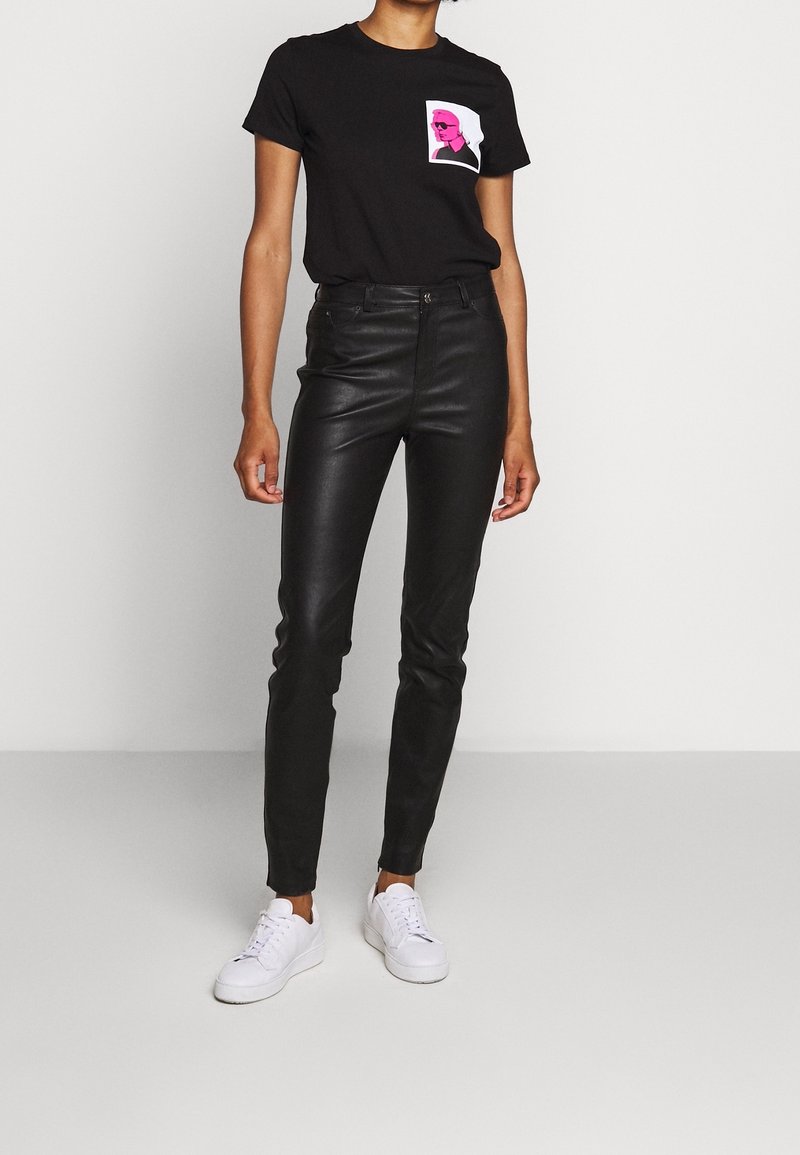 Persona che indossa una maglietta nera con grafica rosa, pantaloni in pelle nera e sneaker bianche, in piedi contro uno sfondo chiaro e uniforme.