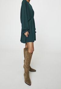 Robe texturée teal avec des manches longues et une taille froncée, associée à de grandes bottes de cowboy en daim marron avec des coutures décoratives.