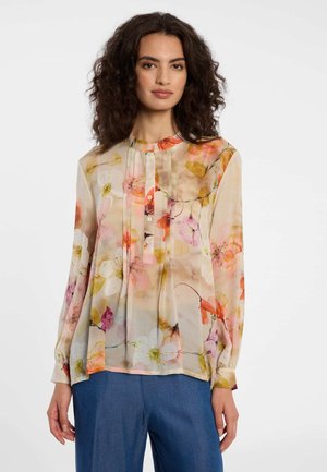 Femme portant un chemisier transparent à manches longues avec un imprimé floral rose, orange et blanc, associé à un pantalon bleu ample sur un fond blanc.