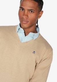 Beiger V-Ausschnitt-Pullover aus weichem Strickmaterial, getragen über einem hellblauen und weißen, gestreiften Hemd mit Knöpfen. Verfügt über ein kleines blaues Logo auf der Brust.