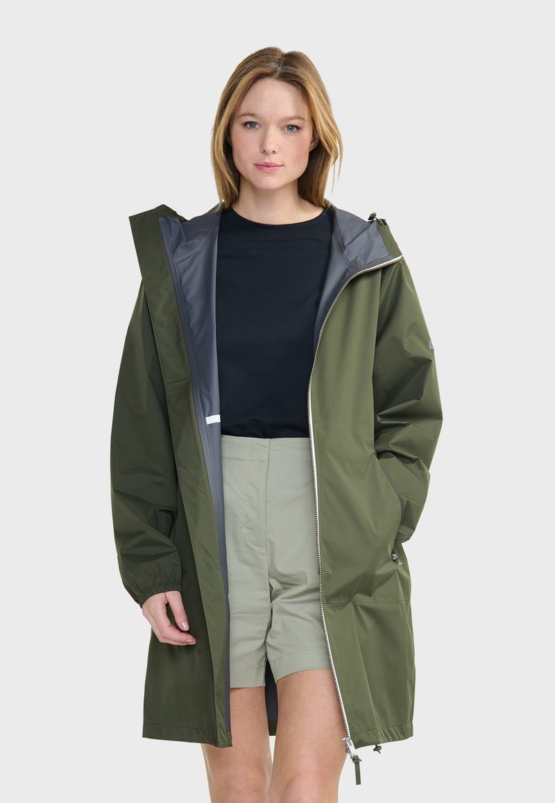 Veste de pluie vert olive avec un intérieur gris, fermeture éclair à l'avant, poignets élastiques et poches latérales. Portée par-dessus une chemise noire et un short vert clair.