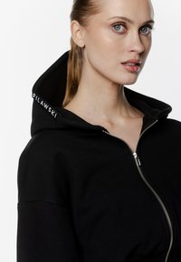 Czarna bluza z kapturem na zamek błyskawiczny i wysokim kołnierzem, z srebrnym zamkiem oraz wyszywanym logo marki na kapturze. Tkanina wydaje się miękka o gładkiej fakturze.