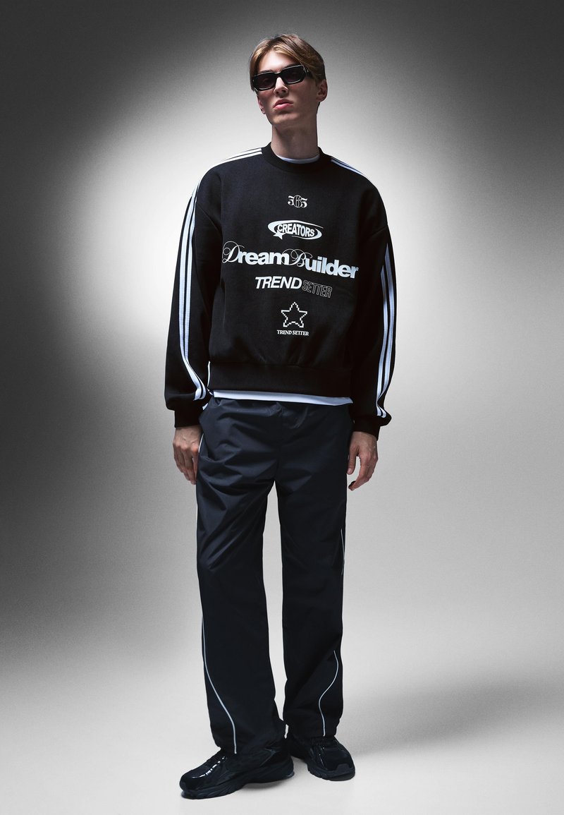 Fekete pulóver fehér grafikákkal és csíkokkal, fekete nadrággal, amelyen fehér szegély található, és fekete sportcipőkkel. A modellen napszemüveg van.