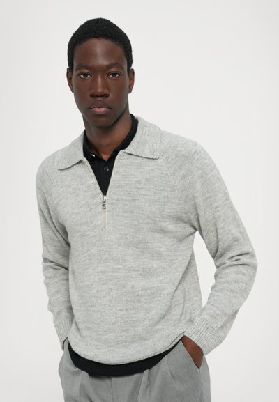Suéter gris claro con cuello con cremallera, puños y dobladillo acanalados. Llevado sobre una camiseta polo negra, que presenta una textura de punto suave.