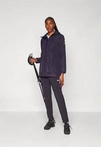 Veste imperméable marine avec capuche, fermeture éclair avant et deux poches, portée avec un pantalon assorti et des chaussures foncées, tenant un parapluie.