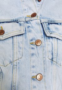 Ljusblå denimjacka med koppartonade knappar, gula sömmar och två bröstfickor. Distressed-textur ger extra detalj.