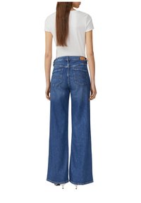 Jean en denim bleu à taille haute et jambes larges, avec deux poches arrière et des détails de couture visibles. Porté avec un haut blanc à manches courtes.