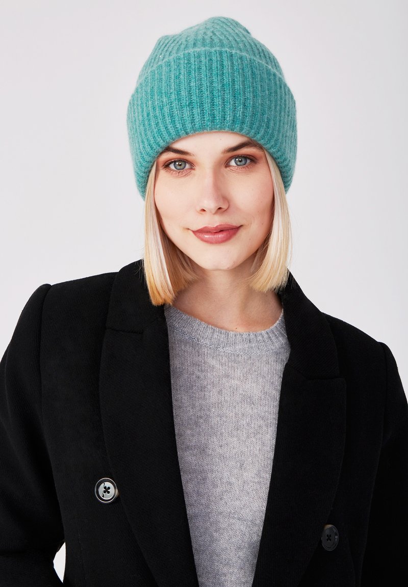 Berretto a coste turchese con orlo piegato, indossato con un cappotto nero e un maglione grigio, caratterizzato da una trama liscia e una vestibilità aderente.