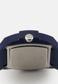 Montre-bracelet en silicone bleue avec un dos en acier inoxydable, un boîtier rond et une couronne argentée proéminente. Comprend le logo de la marque gravé à l'arrière.