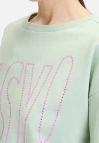 Vaaleanvihreä collegepaita, jossa on ribbauskaulus ja suuria vaaleanpunaisia kirjainkoristeita, joissa on pieniä strasseja.