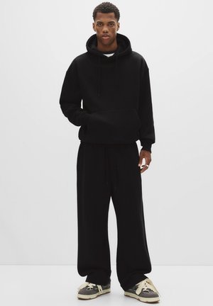Sort hættetrøje med kængurulomme, parret med løstsiddende sorte sweatpants. Modellen har grå sneakers på med beige snørebånd og detaljer.