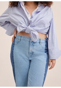 Femme portant une chemise rayée bleu clair nouée à la taille et un jean bicolore bleu, main posée sur la hanche, fond neutre.