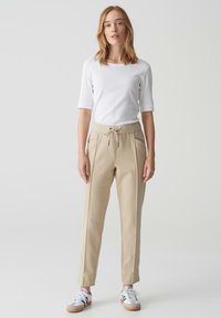 Beige tapschoenen met een zachte textuur en een trekkoord in de taille. Gekleed met een wit T-shirt met korte mouwen en sneakers met grijze en blauwe accenten.