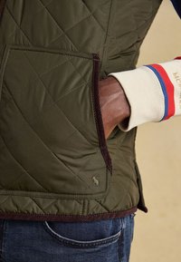 Hand in der Tasche einer olivgrünen gesteppte Jacke mit braunem Besatz, trägt einen cremefarbenen Ärmel mit roten und blauen Streifen und dunkelblaue Jeans.