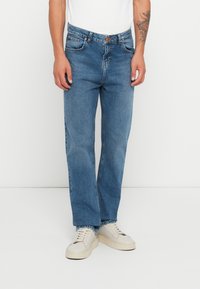 Vaqueros de denim azul claro con corte recto, dobladillo sin rematar, diseño de cinco bolsillos, y detalles en cobre, combinados con zapatillas crema.