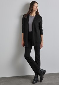 Svart stickad cardigan över en grå topp, i kombination med svarta byxor och svarta kängor i ankellängd. Slät textur och öppen framsidan design.