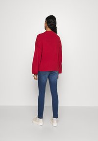 Czerwony, oversize sweter z długimi rękawami, w połączeniu z dopasowanymi niebieskimi dżinsami. Znaczące detale to zaokrąglony dekolt i casualowe obuwie.