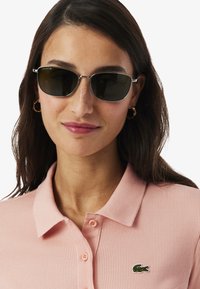 Occhiali da sole con lenti scure, montatura in metallo argento, indossati con una polo rosa chiaro a coste con un piccolo logo Lacoste verde.