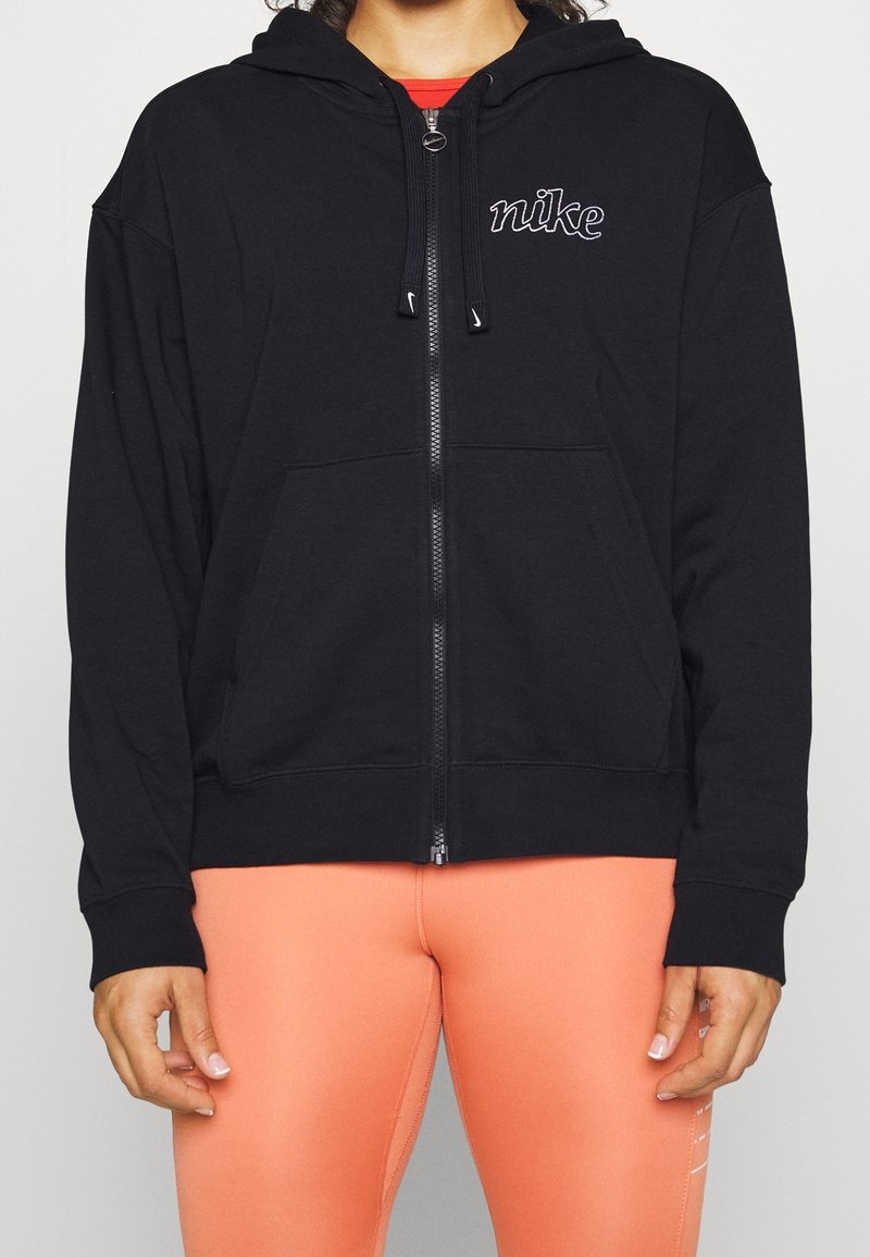 Personne portant un sweat à capuche noir Nike zippé avec une capuche et des poches à l'avant, associé à des leggings orange vif.