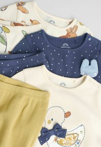 Samling av babytillbehör som inkluderar en krämfärgad topp med anka broderad och rosett, en marinblå topp med vita prickar, och ett par senapsgula byxor.