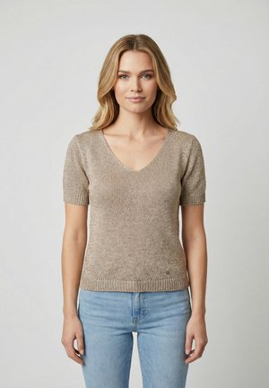 Donna con capelli biondi che indossa un top beige a maniche corte in maglia e jeans azzurri, in piedi su uno sfondo chiaro e uniforme.