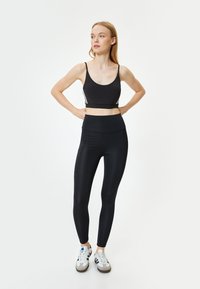 Zwarte sportbeha met dunne bandjes, gecombineerd met hoge zwarte leggings. De leggings hebben een gladde textuur en zijzakken.