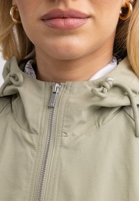 Svetlo zelena jakna z zadrgo spredaj, prilagodljivimi vrvicami na kapuci in gladko teksturo. Znotraj ima vzorčasto podlogo.