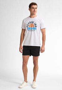 Tricou alb din bumbac cu grafică "PETROLIND" care prezintă un soare și valuri în nuanțe de portocaliu și albastru. Purtați cu pantaloni scurți negri și adidași deschiși.