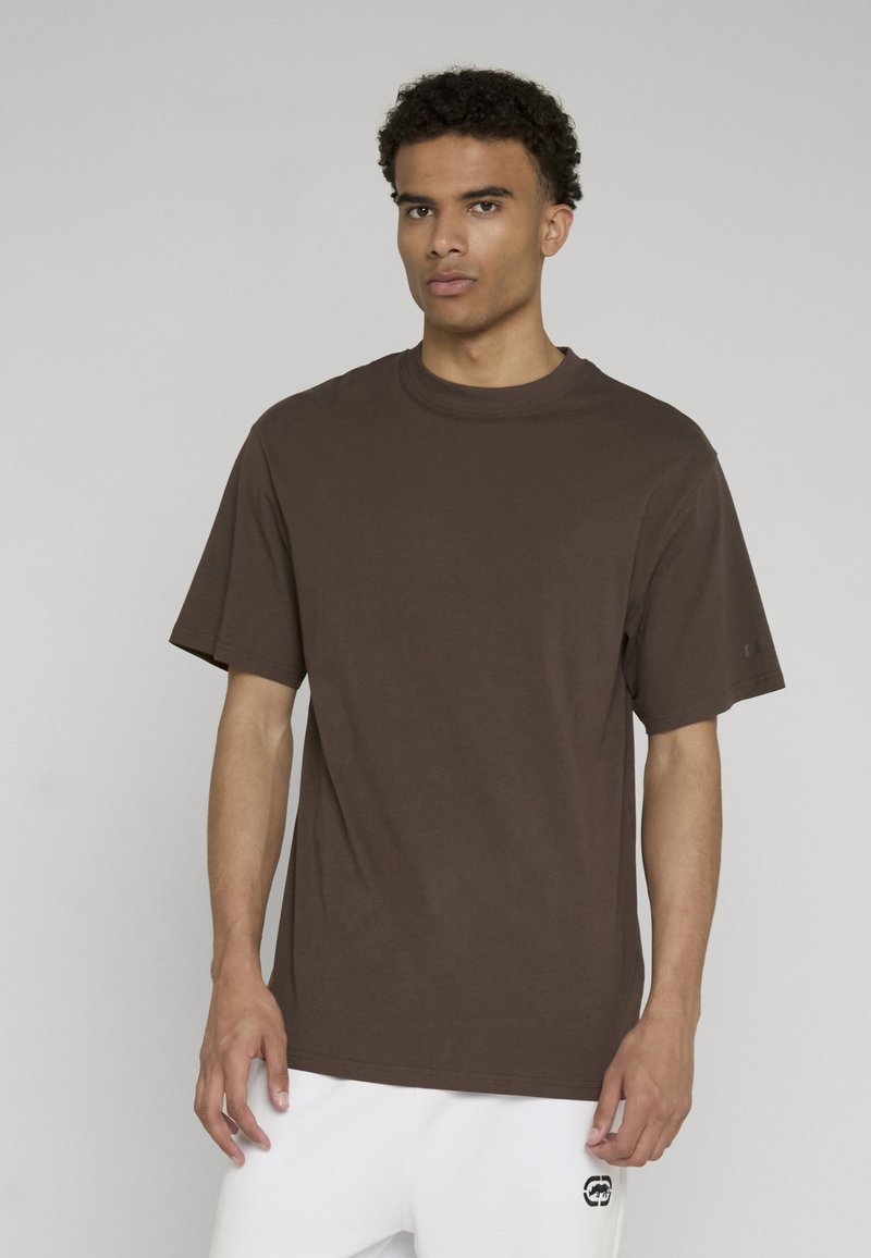 Jeune homme aux cheveux bouclés portant un t-shirt uni marron foncé à manches courtes et un pantalon blanc, debout devant un fond uni.