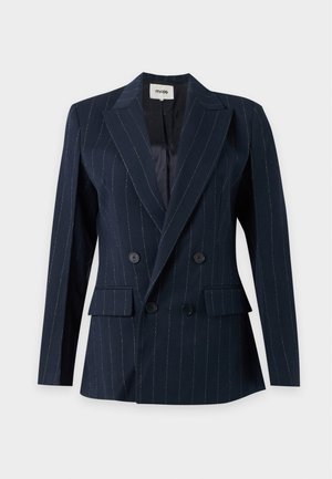 Donna che indossa un blazer doppiopetto a righe scure, camicia bianca con colletto, jeans azzurri chiari e tacchi bianchi a punta, in piedi davanti a uno sfondo semplice.