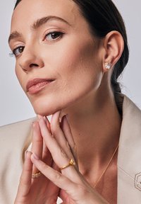 Złote kolczyki z przezroczystym kamieniem, złoty pierścionek z przezroczystym kamieniem oraz delikatny złoty naszyjnik na neutralnym tle.