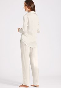 Ensemble de pyjama en satin couleur crème clair, comprenant un haut à manches longues avec des boutons, à ourlet arrondi, et un pantalon droit. Texture douce, sans motifs.