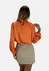 Satijnen, oranje blouse met lange, opgeblazen mouwen en knoopsluiting aan de achterkant. Gecombineerd met een neutrale, aansluitende miniskirt.