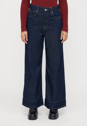 Mørkeblå vide jeans med opsmøgede ben, iført sorte støvler og en langærmet ribbet rød top.