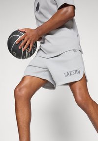 Sive atletske kratke hlače i odgovarajući majica od teksturirane tkanine, s izvezenim natpisom "LAKERS" na hlačama, držeći košarkašku loptu.