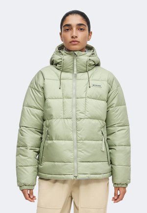 Person trägt eine hellgrüne Columbia Steppjacke mit Kapuze und beige Hose, steht frontal vor einem einfarbigen Hintergrund.