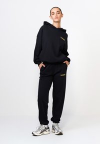 Čierny hoodie a súprava teplákov vyrobená z mäkkej tkaniny, s nápisom "L.A. Minta" v žltej farbe. Nastaviteľná kapucňa, voľný strih a bočné vrecká. Strieborné tenisky.